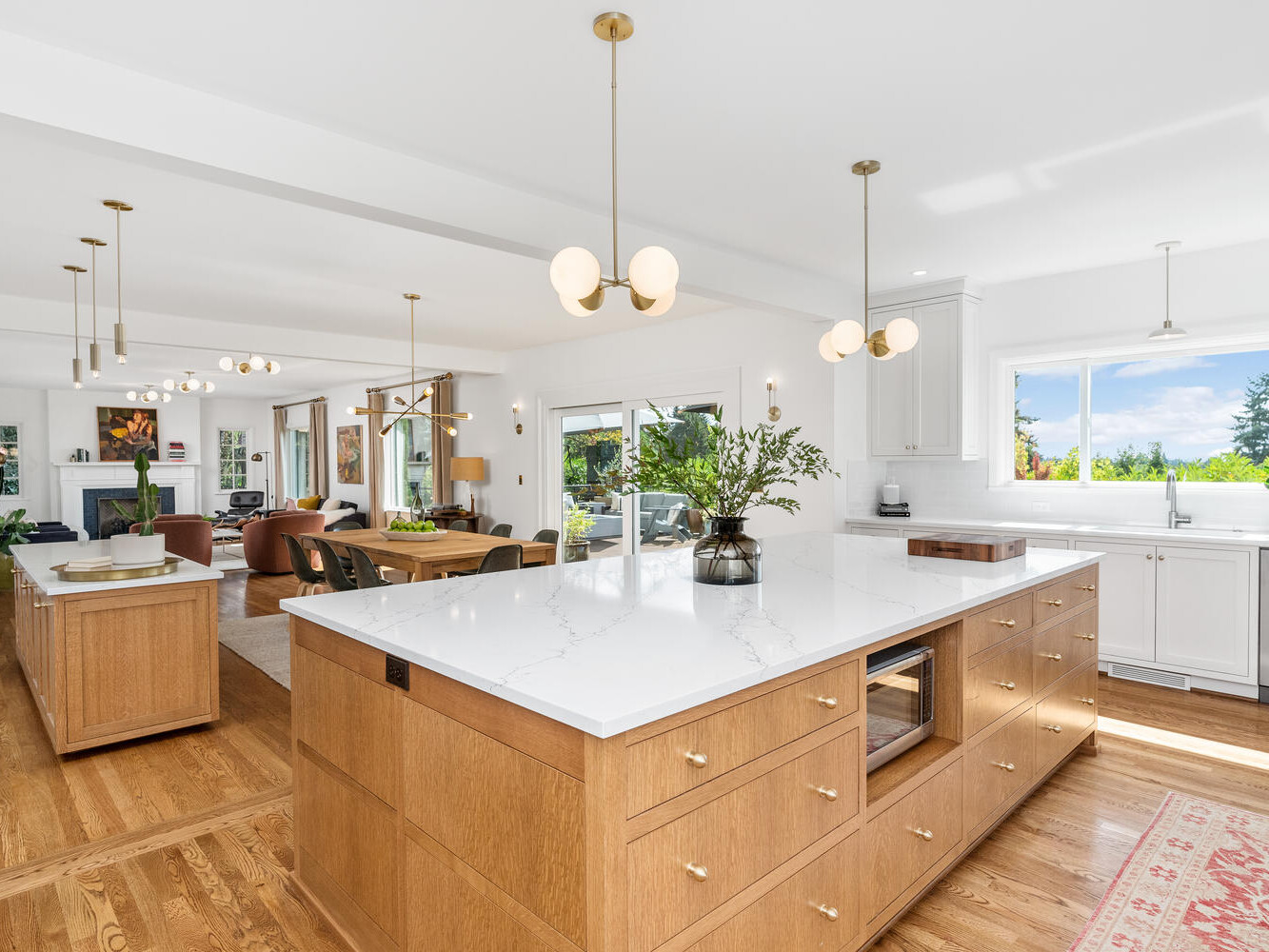 A spacious, modern kitchen in Portland, Oregon features wooden cabinets and a large white marble island. Pendant lights hang from the ceiling, and natural light pours in from large windows. A dining and living area is visible in the background.
