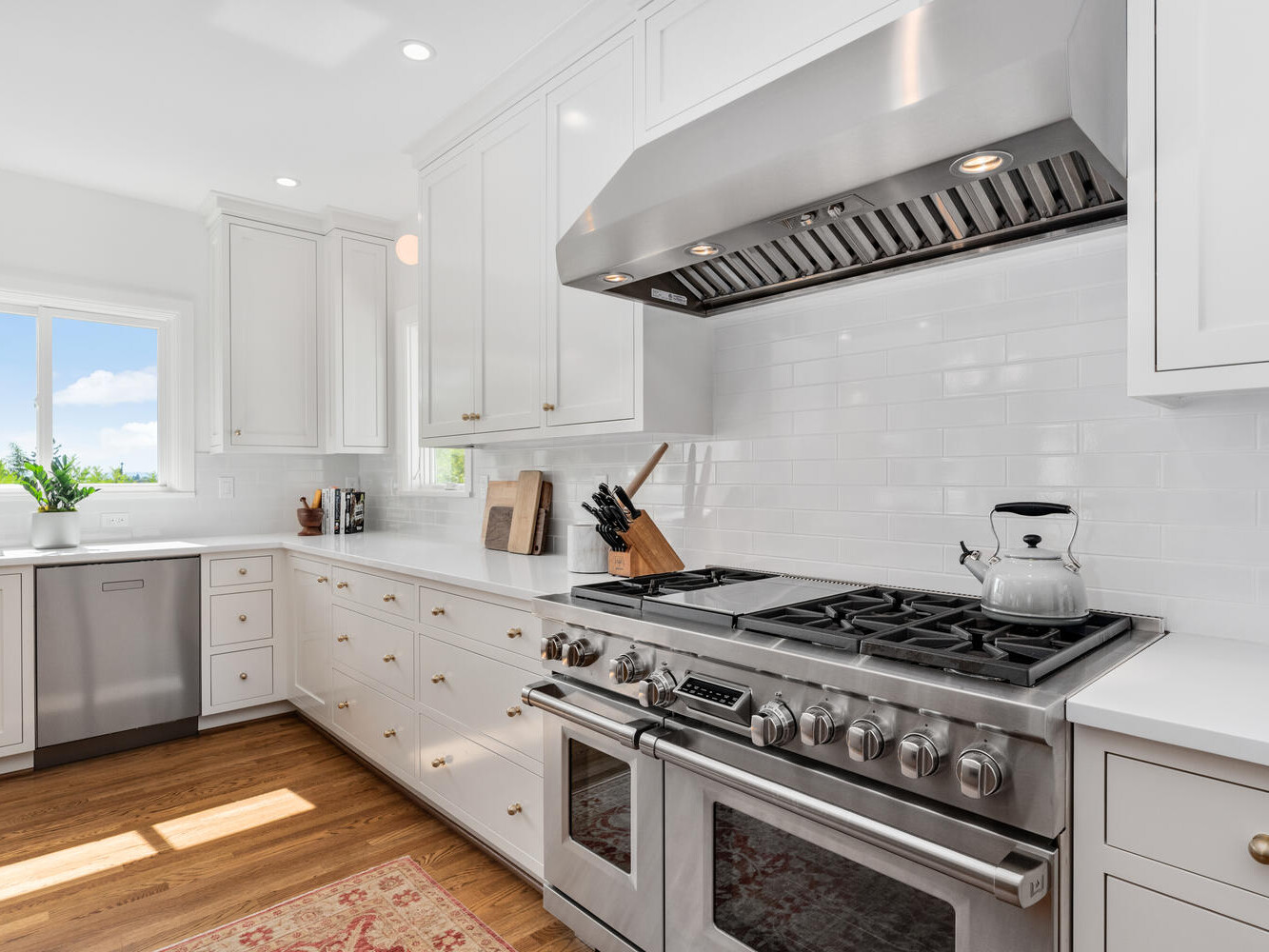 A bright, modern kitchen in Portland, Oregon features white cabinetry and stainless steel appliances, including a large stove with a kettle. Natural light streams through the window, illuminating the potted plant on the counter and wooden floors.