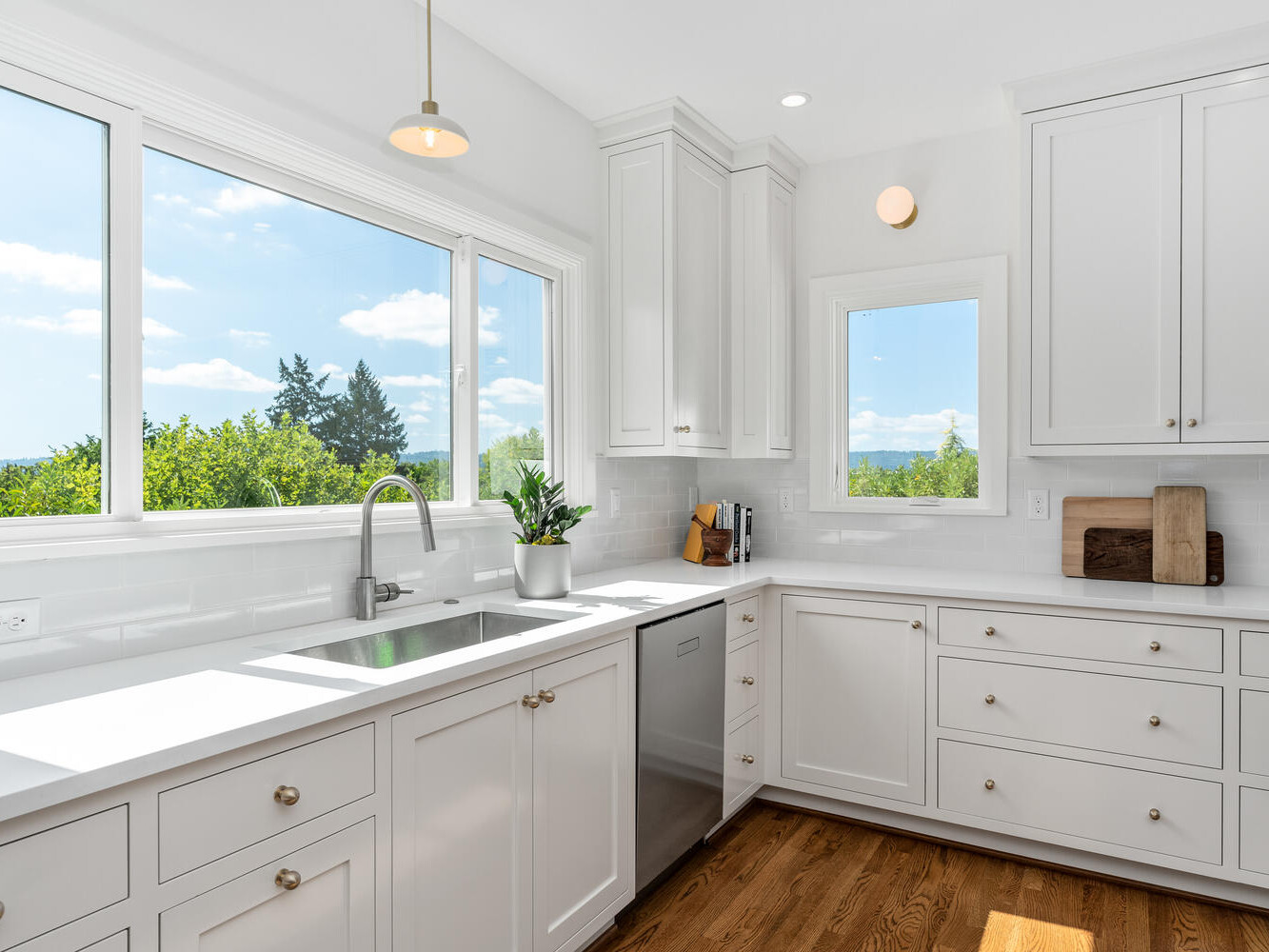 Bright kitchen in Portland, Oregon, with large windows offering a scenic view of trees and sky. The space has white cabinets, drawers, and a sleek countertop featuring a stainless steel sink and dishwasher. A small plant is placed near the window.