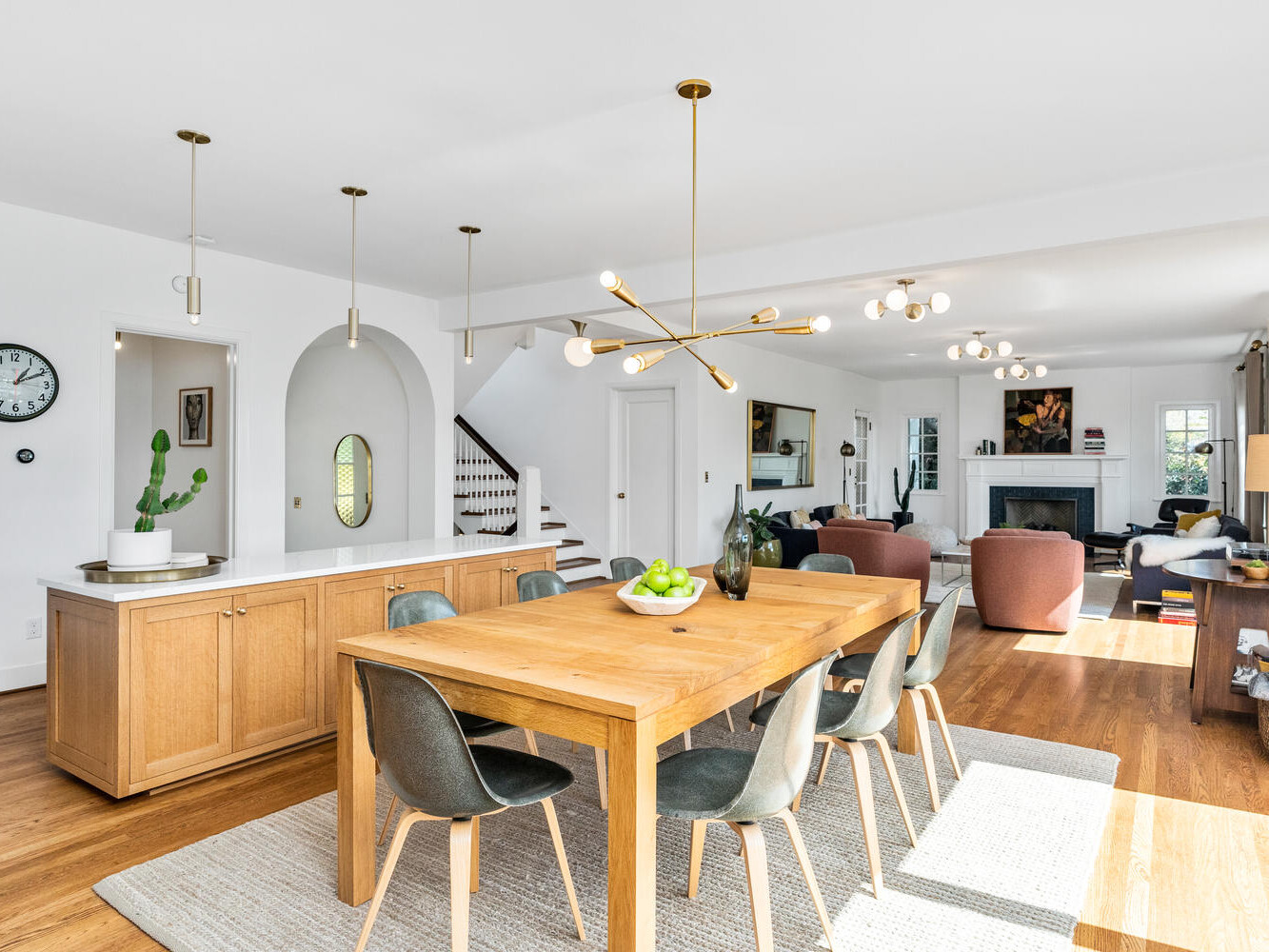 A modern open-concept dining area in Portland, Oregon, features a wooden table with mixed chairs. A kitchen island sits behind with pendant lights above. The space boasts wooden floors, large windows, and a cozy living area with a fireplace in the background.