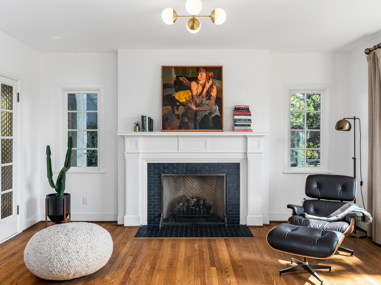 A cozy living room in Portland, Oregon features a black leather lounge chair, an ottoman, and a modern floor lamp. A white fireplace with black tile sits under a framed painting. Nearby, a tall green plant rests by French doors as sunlight filters through the windows.