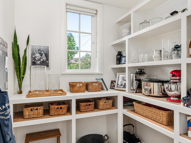 A well-organized pantry in Portland, Oregon, features white shelves holding baskets, kitchen appliances, and jars in a bright room with a wooden floor. A window with a view of trees brings in natural light, while a framed photo decorates the wall.