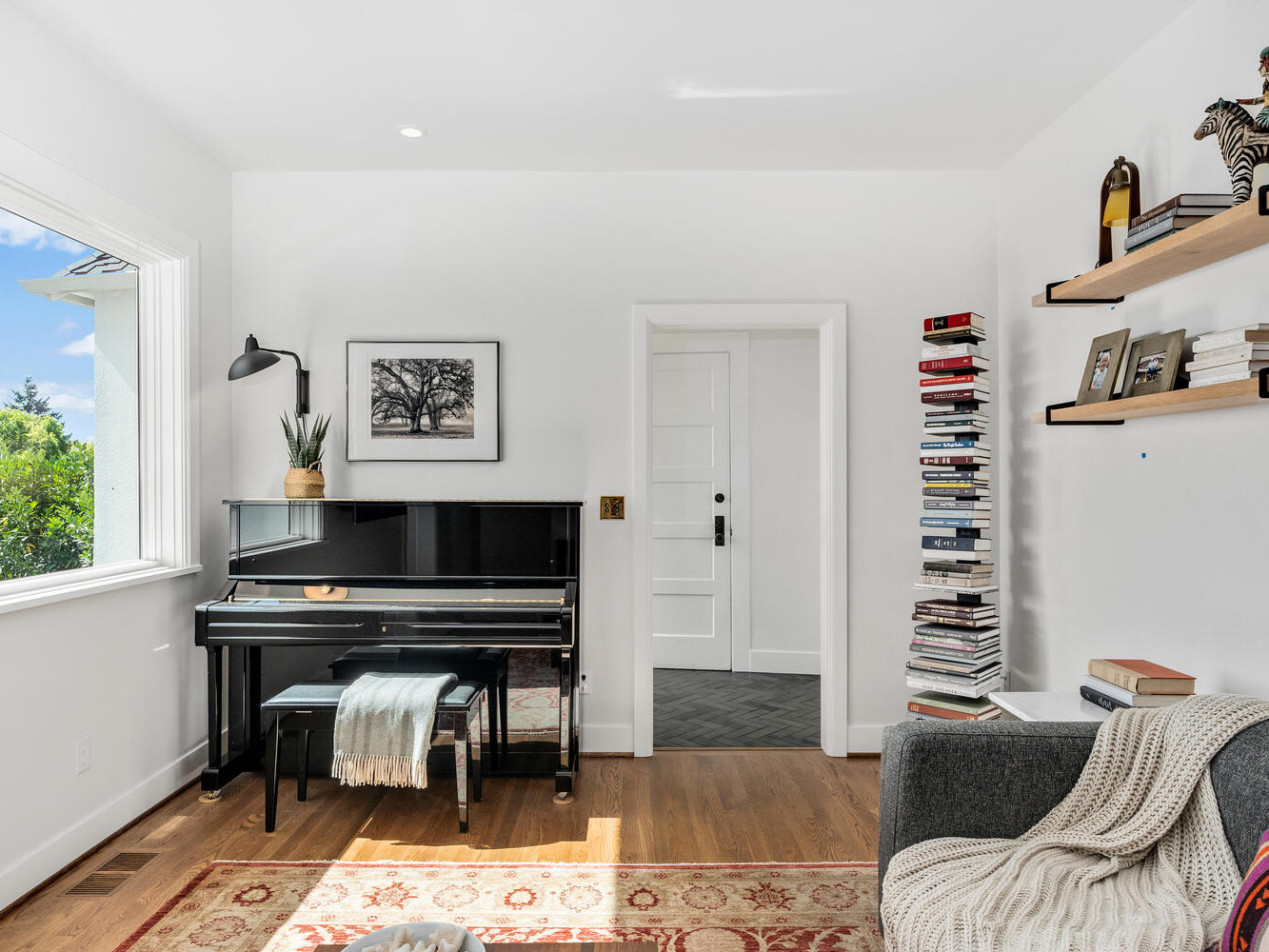 A cozy living room in Portland, Oregon, features a piano, a small bench, and framed art on the wall. A stack of books rests beside the door, with two wooden shelves displaying decorative items above a gray sofa. Sunlight streams in through a large window.