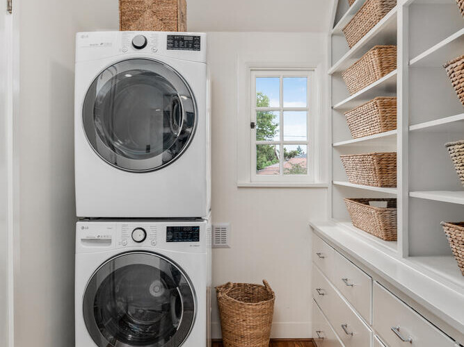 A bright laundry room in Portland, Oregon features a stacked washer and dryer on the left, surrounded by organized wicker baskets on shelves. Natural light spills in through a window, illuminating a beige rug on the wooden floor. White cabinetry lines the right wall.