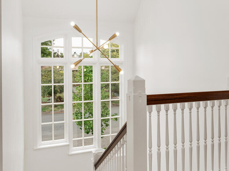 A staircase in a well-lit Portland, Oregon, interior showcases wooden steps and white railings. A modern chandelier hangs from the ceiling, while large windows offer a view of the lush greenery outside.