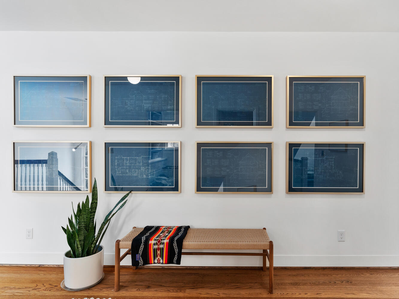 A wall in Portland, Oregon features nine framed blueprints in a grid pattern. Below, a wooden bench with a colorful woven blanket sits next to a potted plant with long green leaves. The wooden floor enhances the minimalist decor.