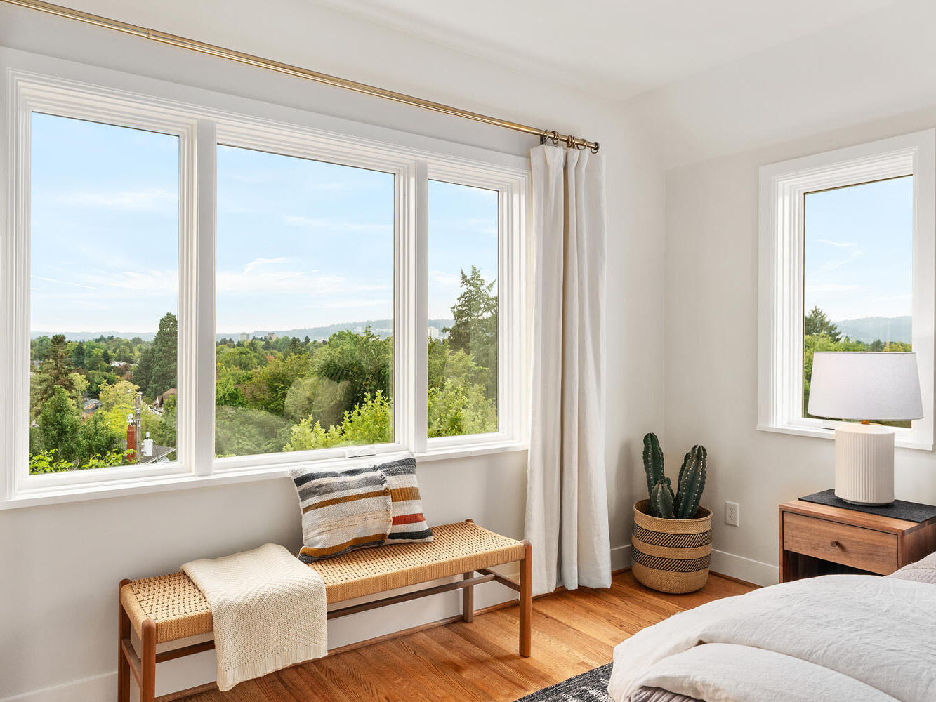 A bright Portland, Oregon bedroom with large windows overlooking a green landscape. A wooden bench with a pillow and throw sits beneath the windows. A potted cactus and lamp are on a small table beside the bed, which is partially visible. Light curtains frame the windows.