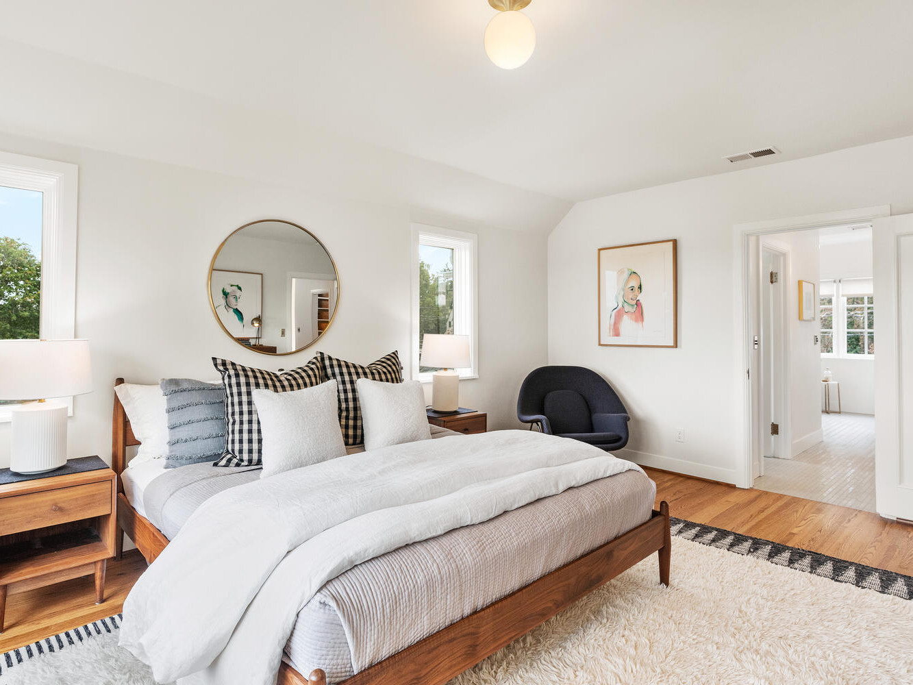 A bright, modern bedroom in Portland, Oregon, features a wooden bed with white and gray bedding and a round mirror above the headboard. Two bedside tables with lamps flank the bed. A dark blue chair sits in the corner beside a doorway leading to a bathroom. Natural light fills the room.