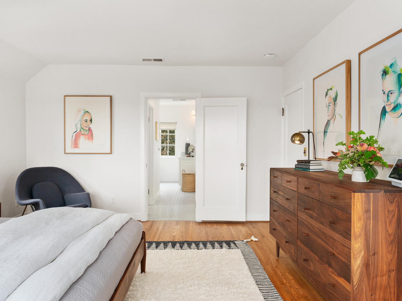 A modern Portland Oregon bedroom with white walls features a wooden dresser, a comfortable bed with light gray bedding, and artwork on the walls. A doorway leads to a bright bathroom, and a dark blue chair sits in the corner. A rug lies on the wooden floor.