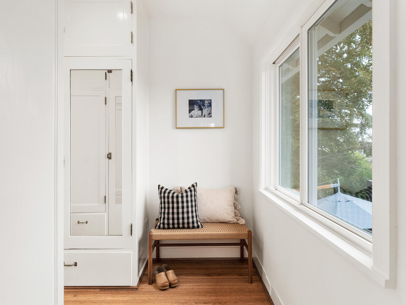 A bright, minimalist hallway in a Portland, Oregon home features a small bench, two cushions—one checkered and one plain—and a pair of slippers on the wooden floor. A framed photo adorns the white wall, while large windows reveal the lush greenery outside.