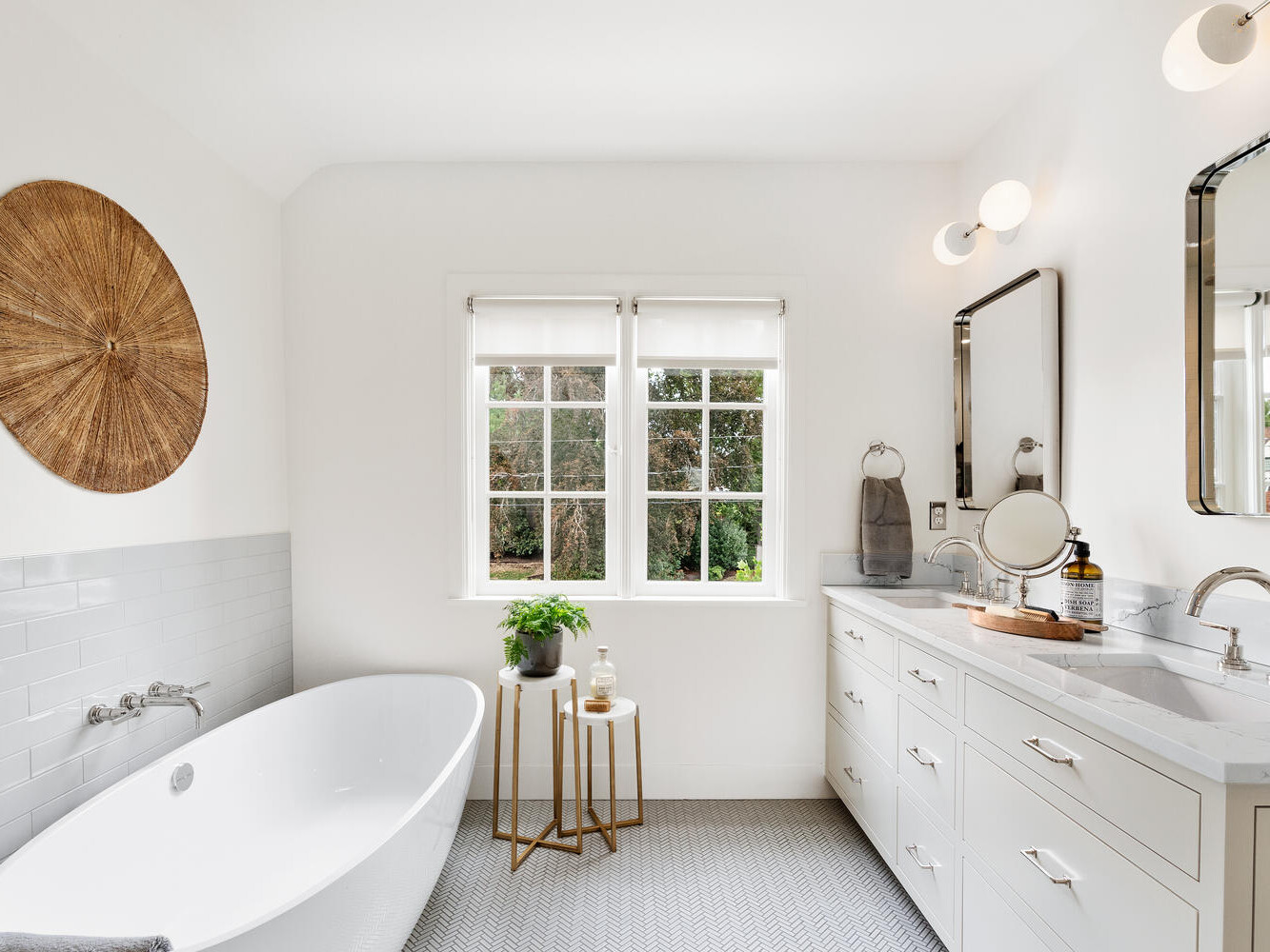 A modern bathroom in Portland, Oregon, features a white freestanding tub and wooden wall decor. Dual sinks with rectangular mirrors reflect the garden view from the window. A small plant on a side table adds life, while soft lighting brightens this serene space.