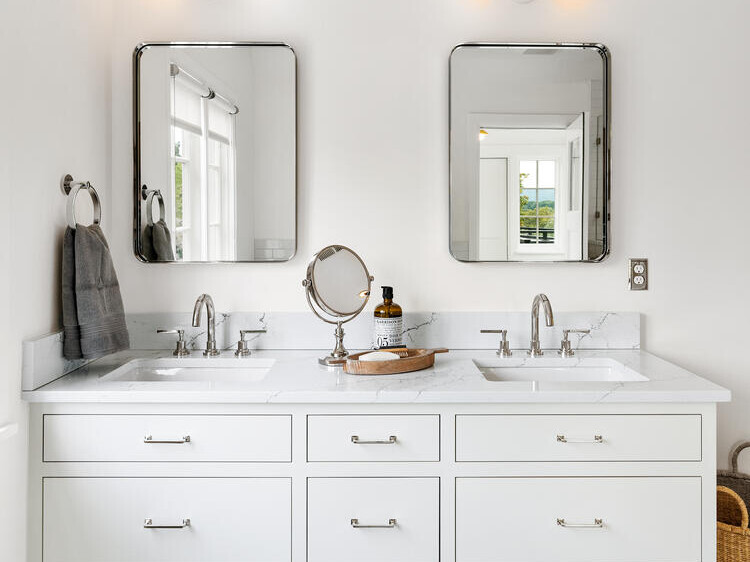 A modern white bathroom in Portland, Oregon features a double vanity, with two rectangular mirrors and lights above. The countertop hosts dual sinks, chrome faucets, a small mirror, and a tray of toiletries. A woven basket neatly rests on the floor.