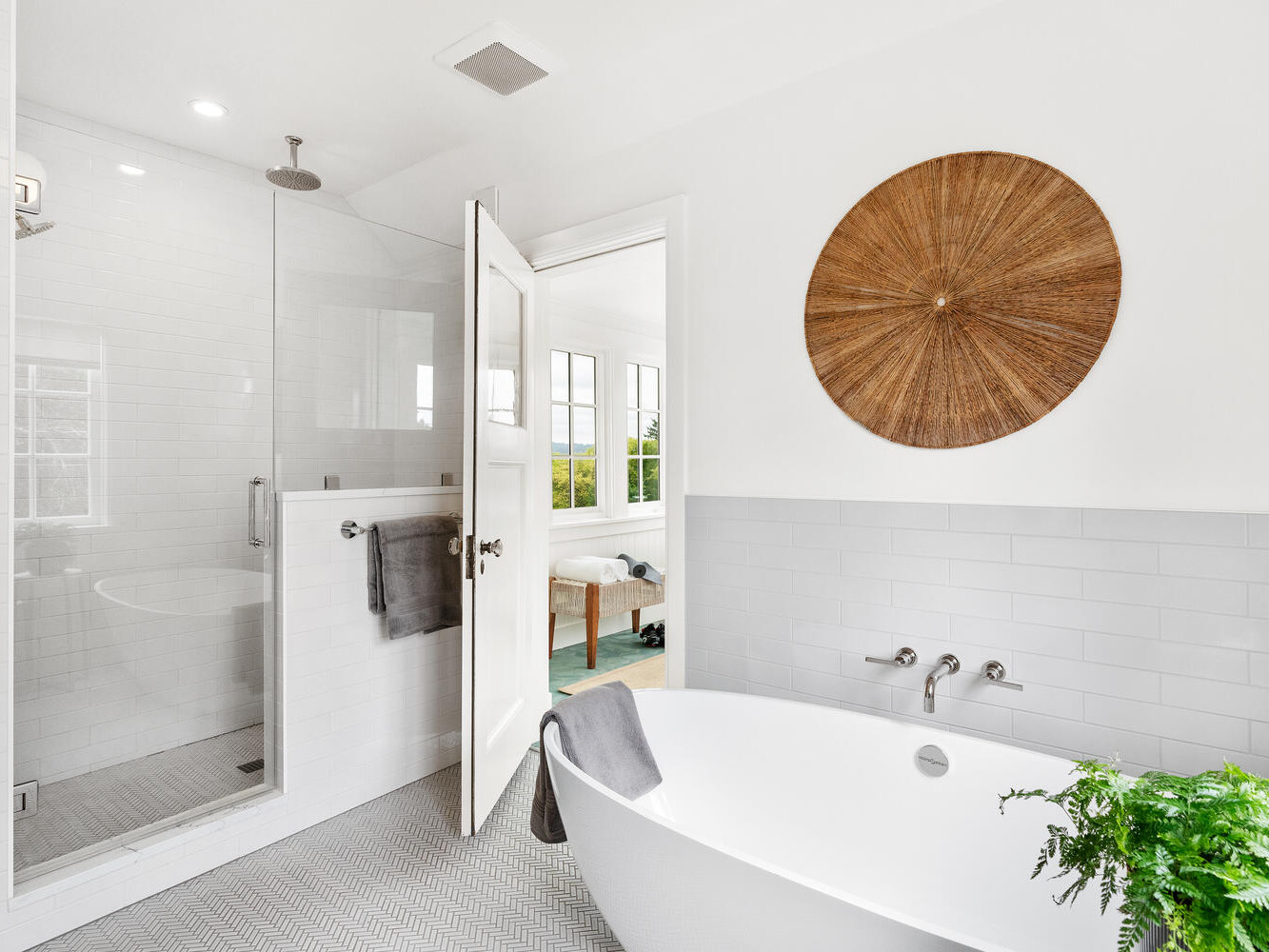 A modern bathroom in Portland, Oregon, features a white freestanding tub, a glass-door shower, and woven circular wall decor. Towels drape artfully on the tub and towel bar, with a plant nearby. Large windows in the adjoining room flood the space with natural light.