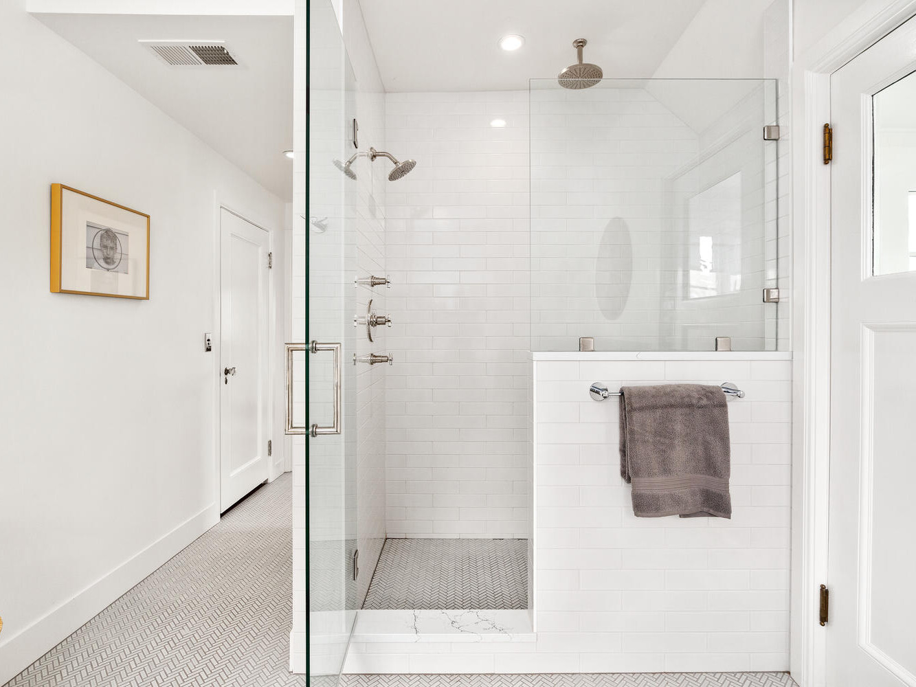Modern bathroom in Portland, Oregon, featuring a glass-enclosed shower with chrome fixtures and a rainfall showerhead. White subway tiles cover the walls, and a gray towel hangs on a rail. A piece of framed art is on the wall, and a woven basket sits on the floor.