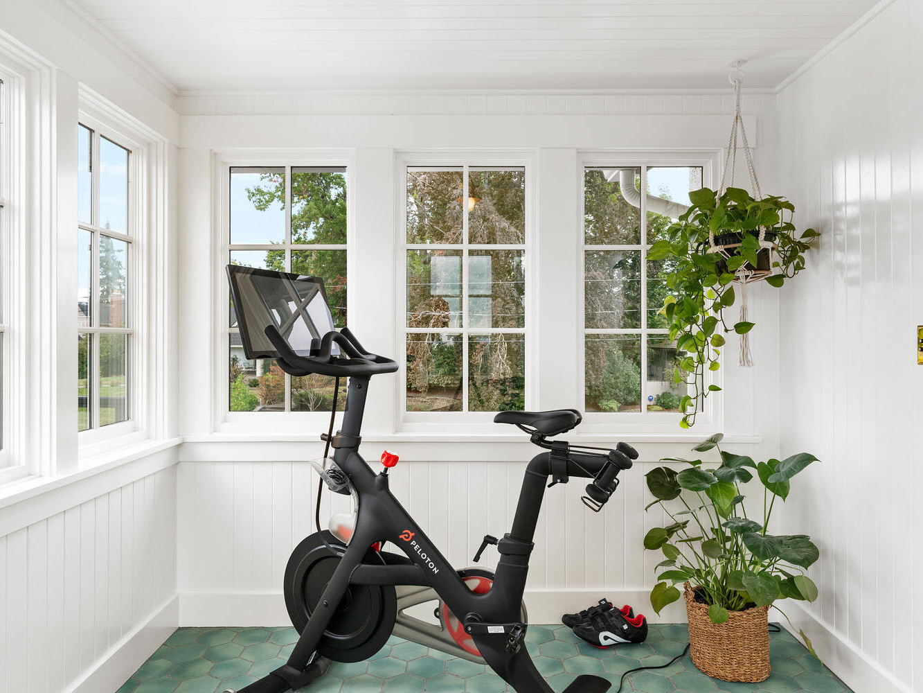 A bright exercise room in Portland, Oregon features a stationary bike positioned in front of large windows. The room boasts white walls, green-tiled flooring, potted plants, and a pair of shoes resting on the floor beside the bike.