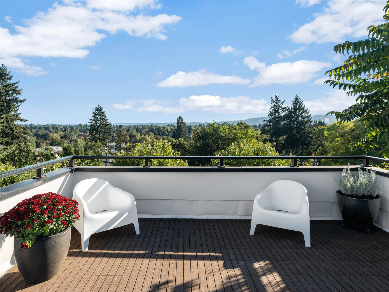 A rooftop patio in Portland, Oregon, features two white chairs and a potted plant with red flowers. The patio overlooks a scenic view of a lush green landscape and distant hills under a partly cloudy blue sky.