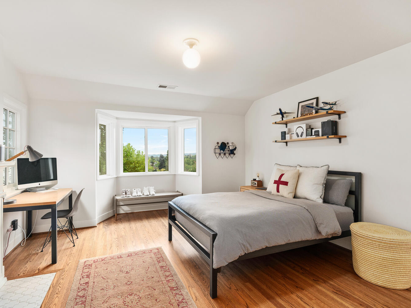 A cozy Portland, Oregon bedroom features a wooden floor, a black metal bed with gray bedding, and a red cross accent pillow. A small desk with a computer sits by the window, offering views of lush greenery. Shelves with decor are above the bed, and a wicker basket is nearby.