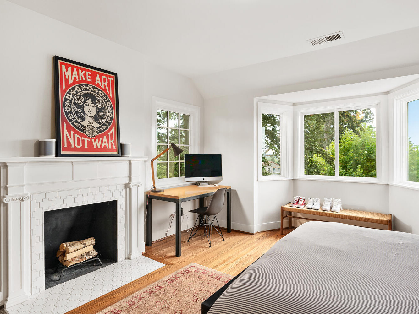 A cozy Portland, Oregon bedroom features a fireplace with a Make Art Not War poster above it. A desk with a computer and lamp sits by a large bay window overlooking greenery. A bed with a gray comforter and a red rug completes this artistic haven.