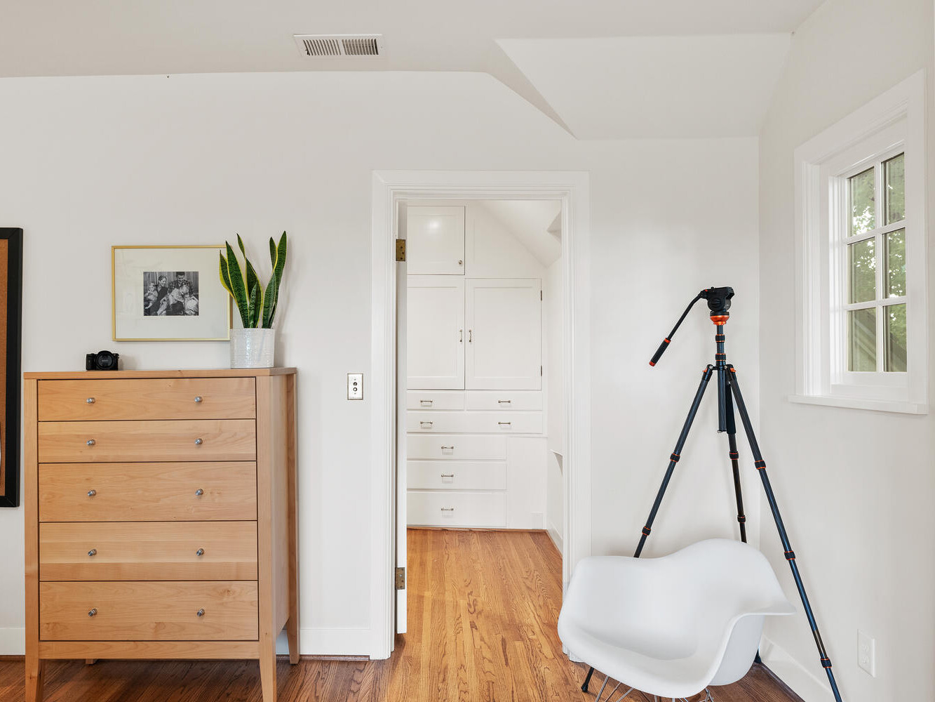 A bright room in Portland, Oregon, features a wooden chest of drawers, a small plant, and pictures on the wall. A camera tripod and a white modern chair are nearby. An open doorway leads to a white closet area with built-in cabinets. Wood flooring throughout.