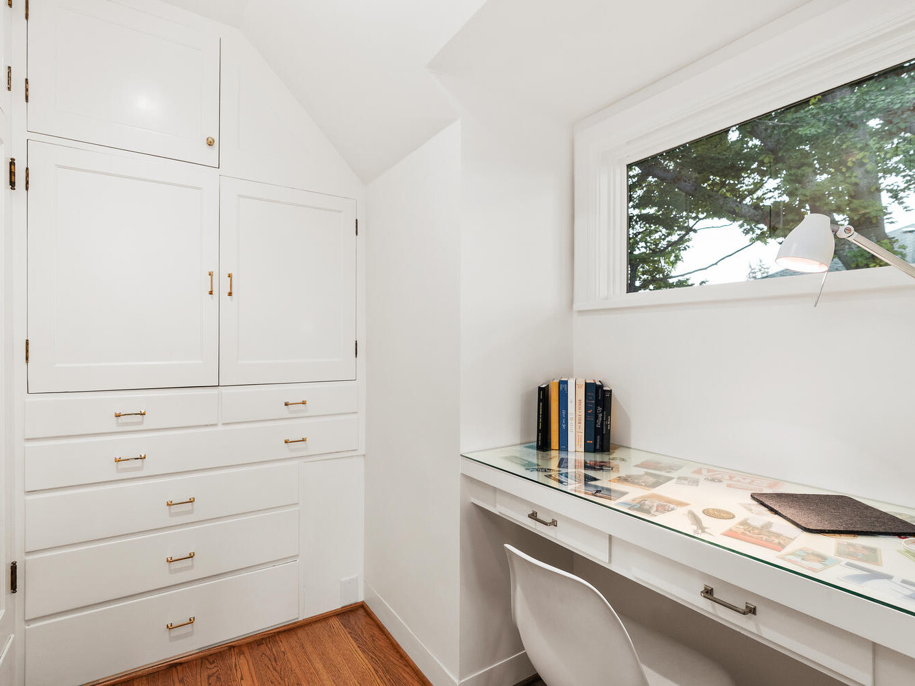 A small, bright home office in Portland, Oregon features a built-in white desk and chair. The desk has a glass surface holding a lamp, books, and mouse pad. A large window lets in natural light, while built-in cupboards with drawers line the left wall.