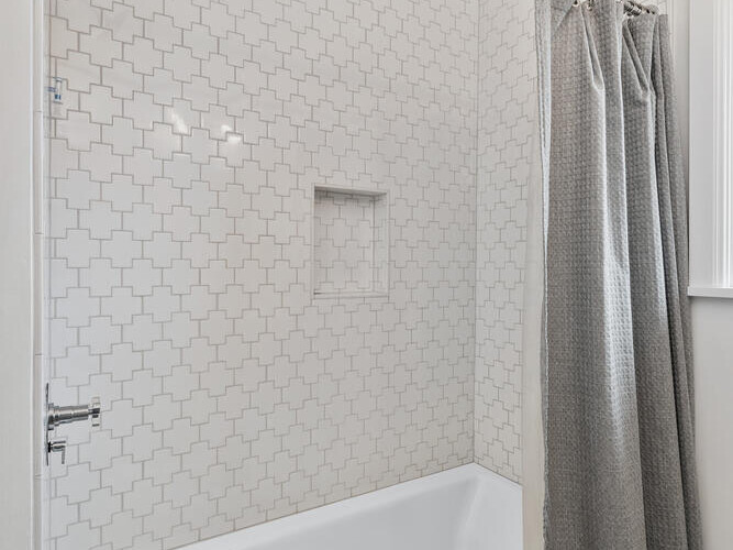 A bathroom in Portland, Oregon, features a white bathtub, patterned wall tiles, and a gray shower curtain. The tile design boasts geometric patterns with a small built-in shelf in the shower area. The floor is elegantly tiled with a small hexagonal design.
