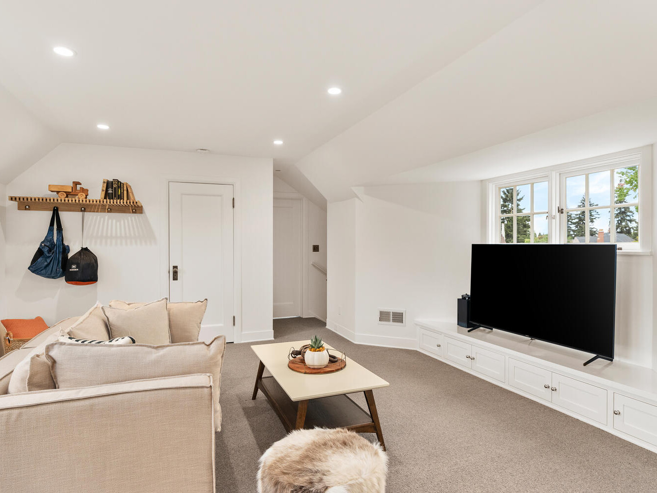 A cozy Portland, Oregon living room features a beige sofa, wooden coffee table, and large flat-screen TV on a white built-in cabinet. The room is well-lit with recessed lighting and natural light from the window. A small shelf with bags and hooks adorns the left wall.