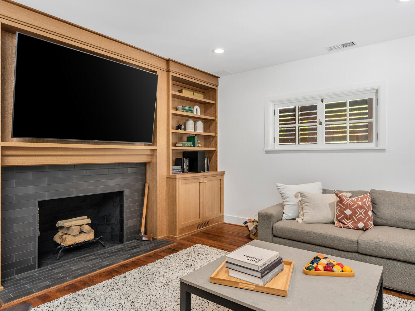 A cozy Portland, Oregon living room features a wall-mounted TV above a fireplace with wood logs. Built-in shelves hold books and decor. A gray sofa with patterned pillows sits opposite a coffee table with magazines and a bowl of colorful balls. A small window provides light.