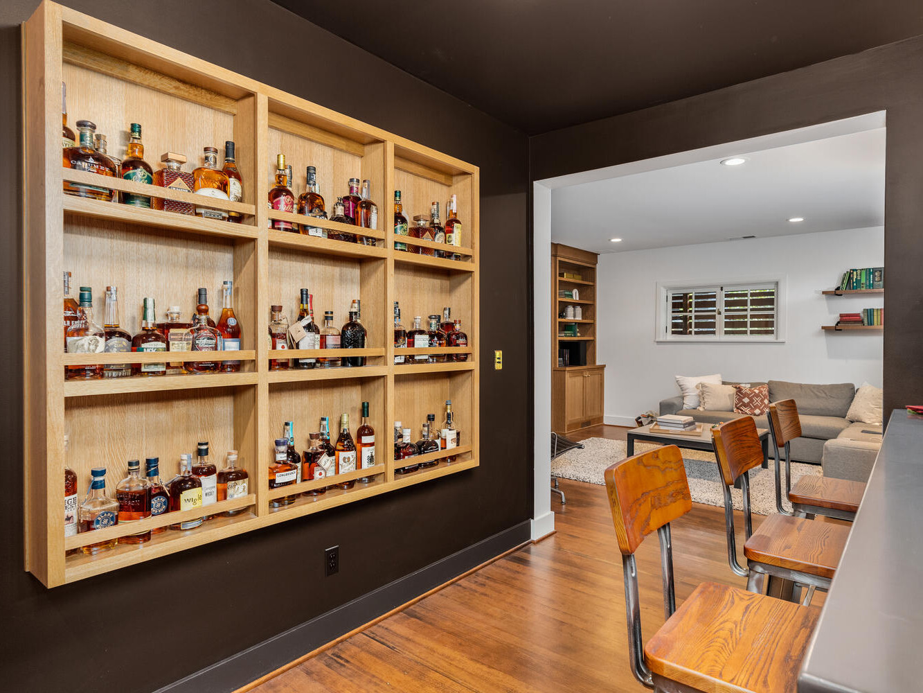 A home bar in Portland, Oregon features a wooden shelving unit displaying assorted liquor bottles. Two wooden bar stools sit at a gray counter. In the background, there's a cozy living area with a couch, a large chair, and a wooden bookshelf.