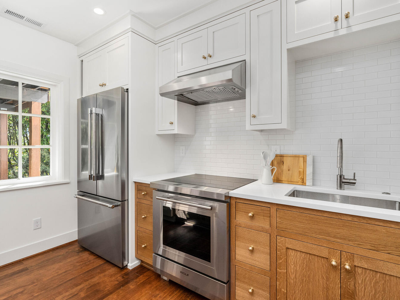 A modern kitchen in Portland, Oregon, with white and wooden lower cabinets features a stainless steel refrigerator, oven, and hood. A white tile backsplash and stainless steel sink are present. Natural light streams through the window, enhancing the contemporary design.