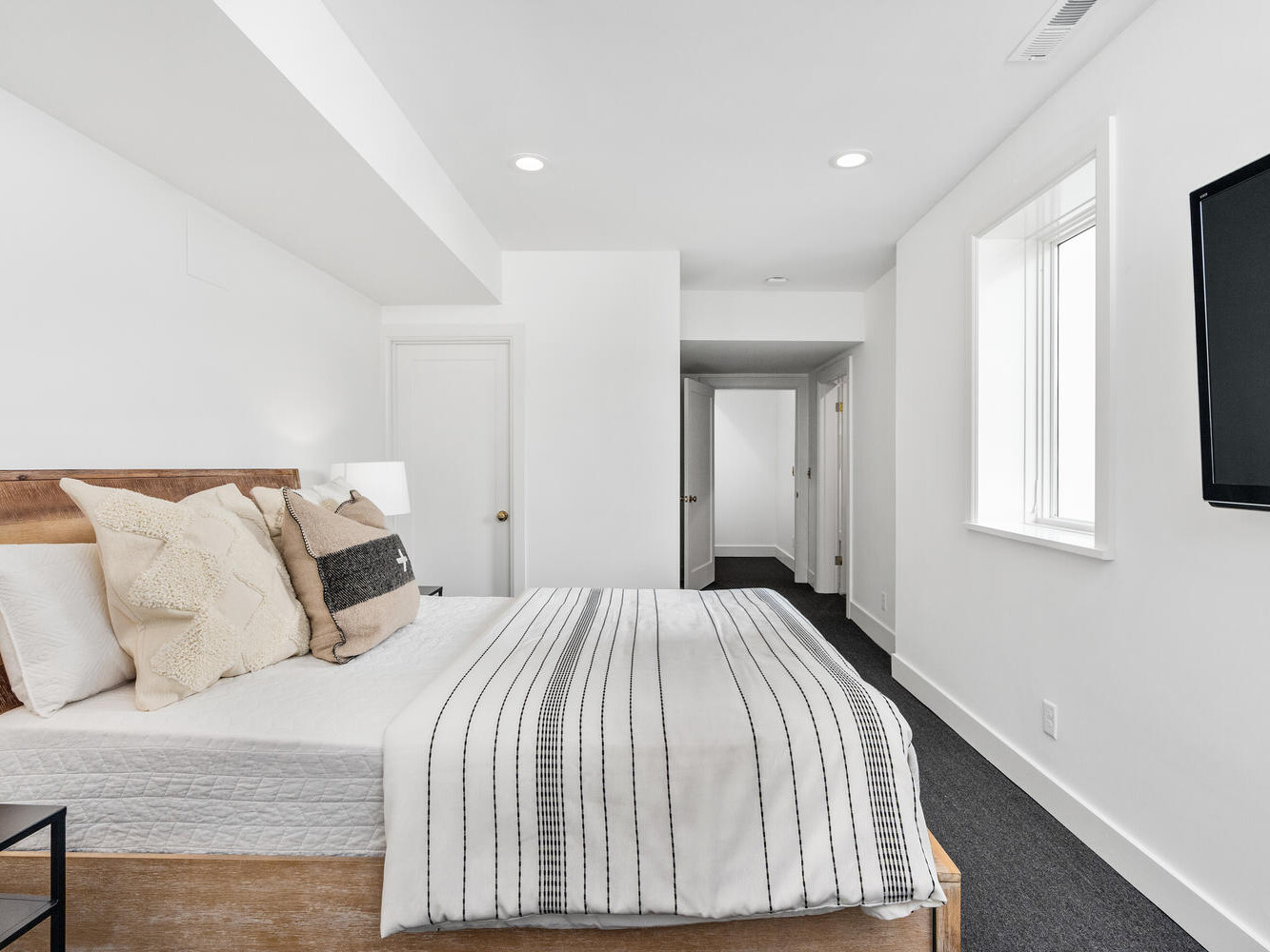 A minimalistic bedroom in Portland, Oregon, boasts a white bed adorned with striped bedding and a mix of white and beige pillows. The room features pristine white walls, a flat-screen TV mounted on the wall, and a dark carpet. Natural light pours in through a window on the right.