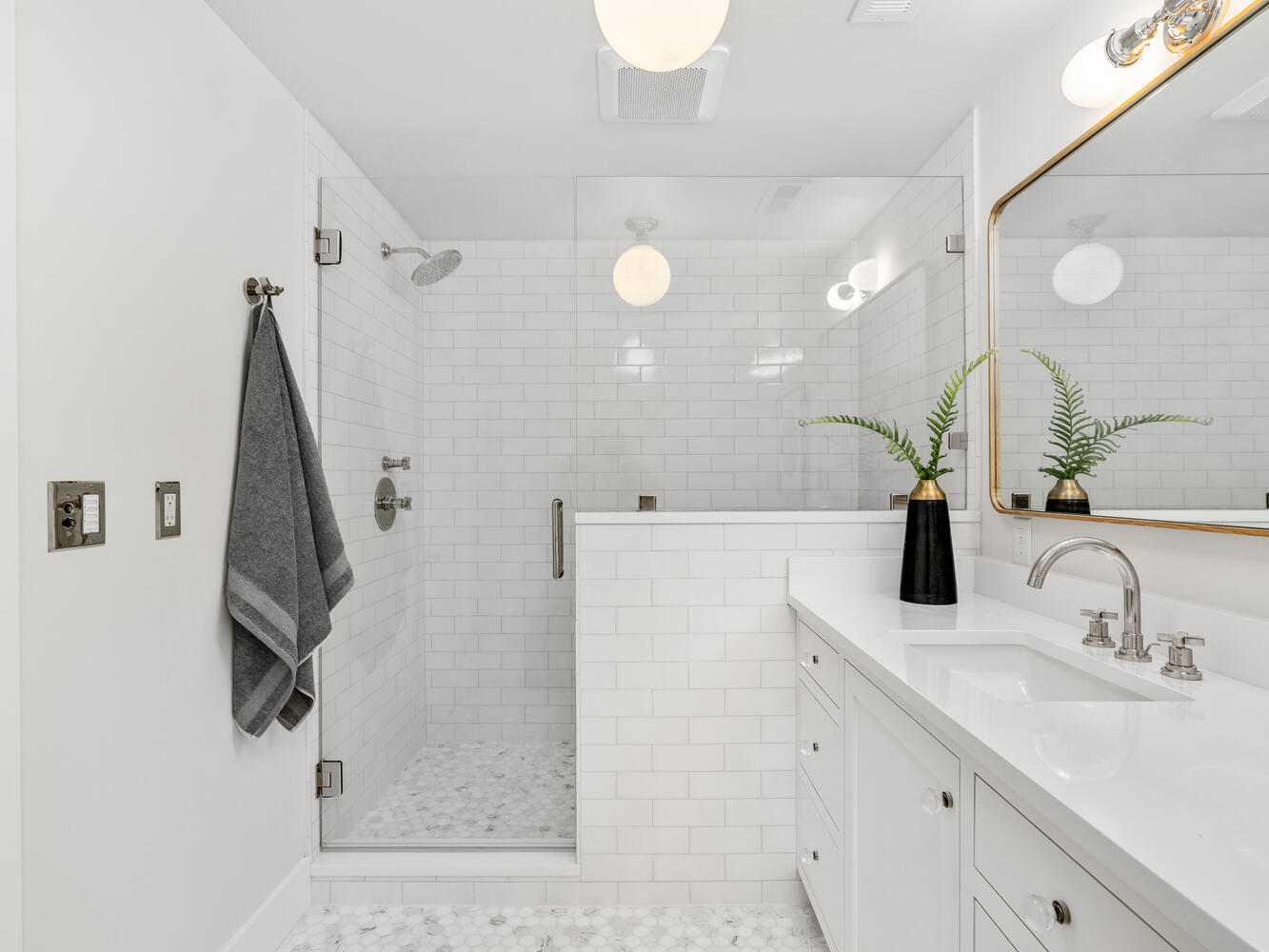 A modern white bathroom in Portland, Oregon, showcases a walk-in shower with a glass door. A dark towel hangs on a silver hook. The vanity features a rectangular mirror, dual sinks, and a black vase with green foliage. Bright, clean tiles adorn the walls and floor.