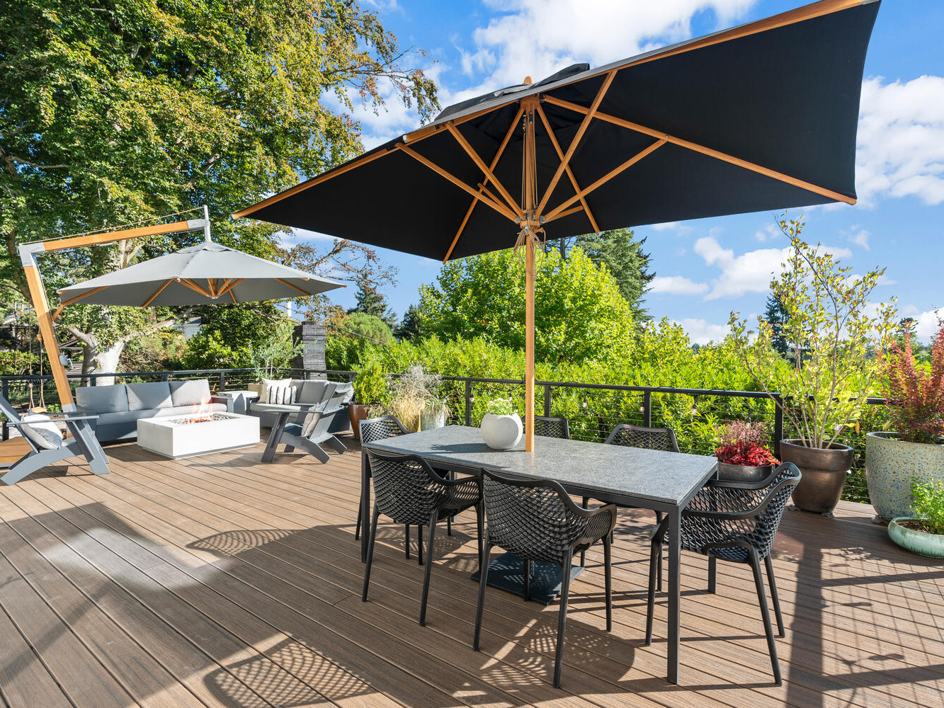 A spacious outdoor deck in Portland, Oregon features a dining table and chairs shaded by large black umbrellas. Lounge chairs and sofas are visible in the background. The deck is surrounded by lush greenery and trees under a bright blue sky.