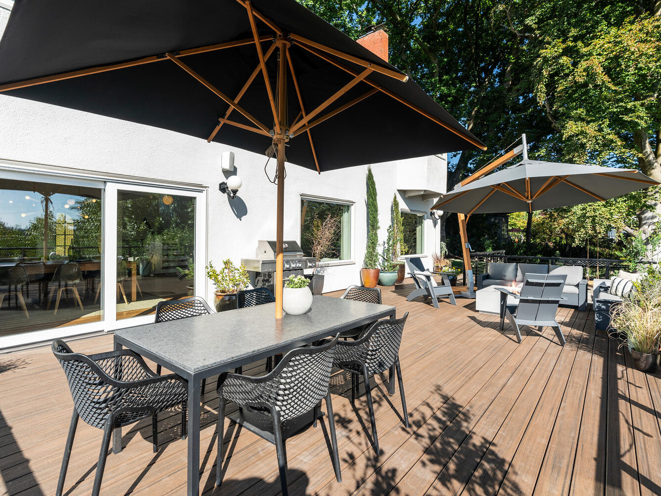 A spacious outdoor deck in Portland, Oregon, features two large black umbrellas shading dining tables and chairs. Sliding glass doors lead to the interior, with potted plants adding charm. Towering trees offer a natural backdrop, crafting a serene atmosphere.