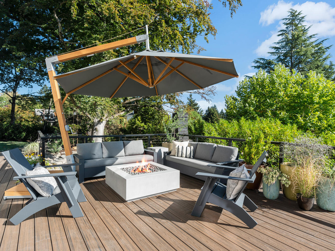 A spacious outdoor patio in Portland, Oregon, features a large umbrella shading gray sofas and chairs around a modern fire pit table. Lush trees and plants surround the wooden deck, creating a relaxing and inviting atmosphere.