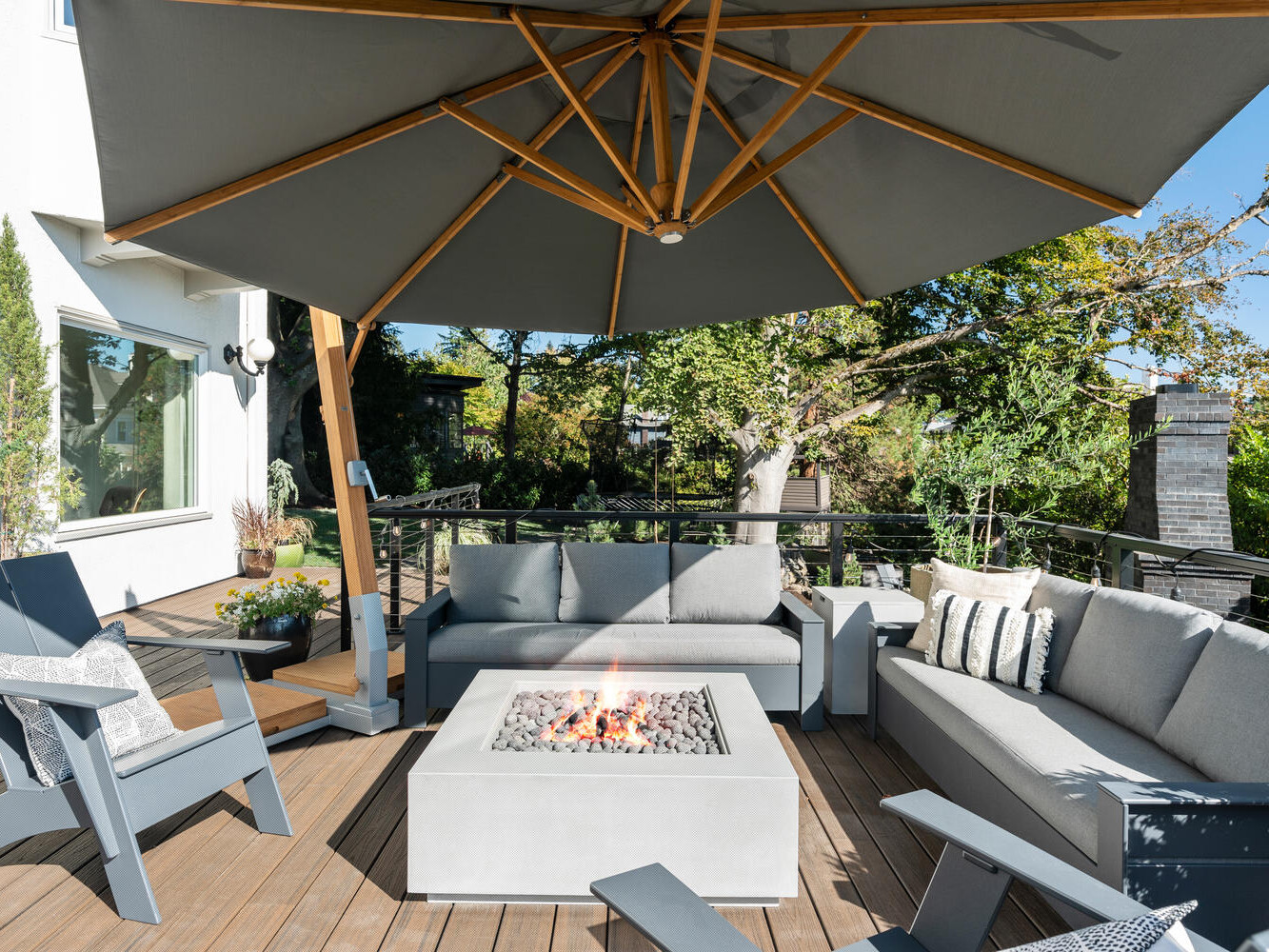 Nestled in Portland, Oregon, this outdoor patio features grey sofas and matching cushioned chairs surrounding a square fire pit. A large grey umbrella offers shade, while the vibrant green trees and plants in the background enhance the serene ambiance.