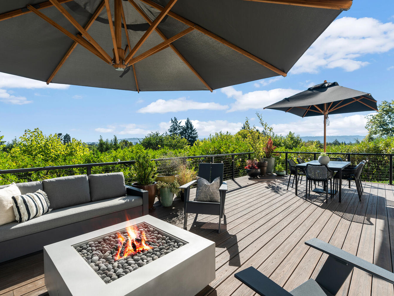 A wooden deck with a fire pit, surrounded by gray seating, overlooks Portland, Oregon's lush green landscape. Two large umbrellas provide shade over the seating area and a dining table with chairs. Blue sky and clouds are visible in the background.