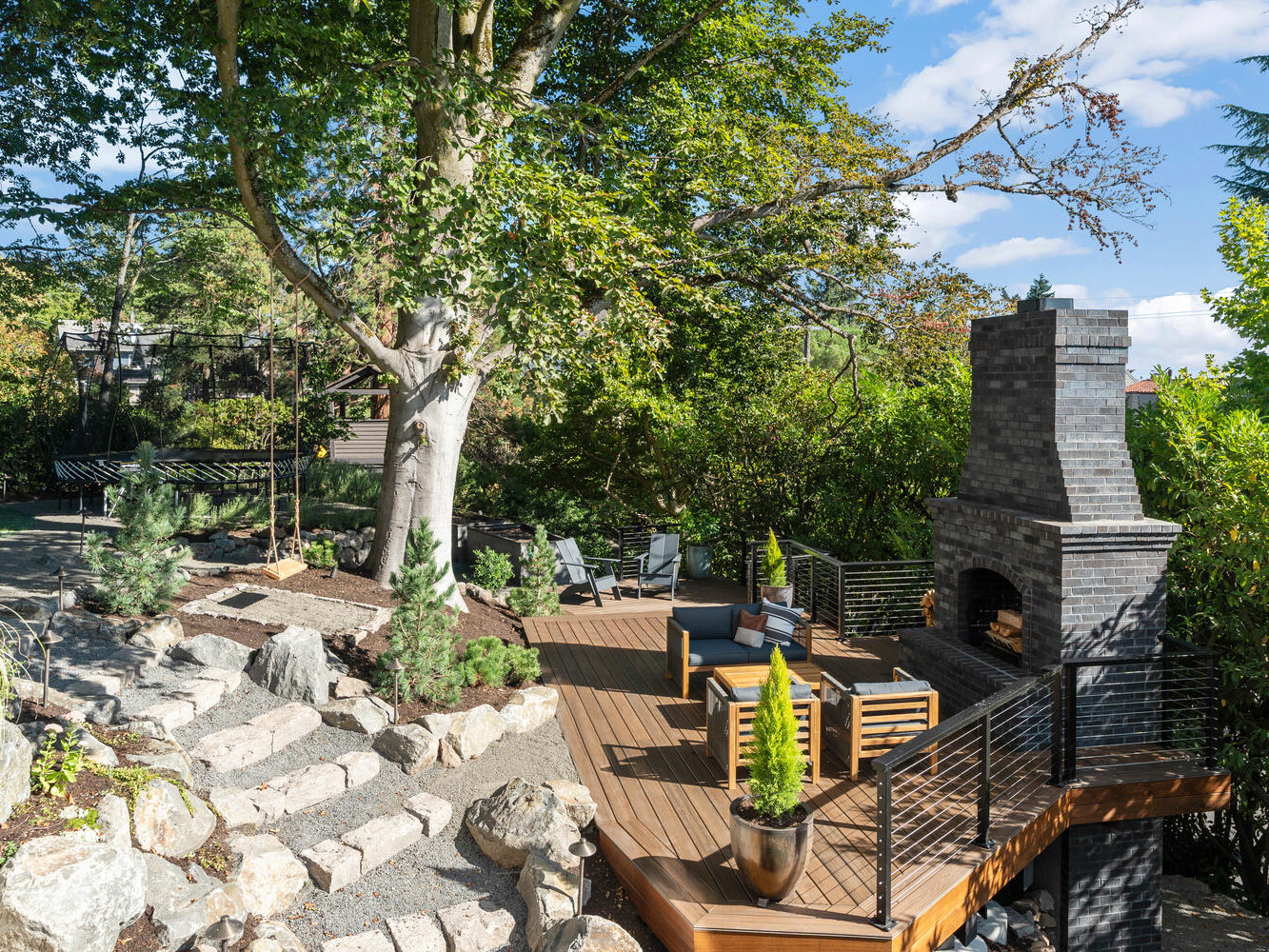 A backyard patio in Portland, Oregon, with a wooden deck surrounded by lush greenery features a tall brick fireplace, modern outdoor furniture, and a winding stone path. A large tree provides shade, enhancing the relaxing, natural atmosphere typical of this Pacific Northwest locale.