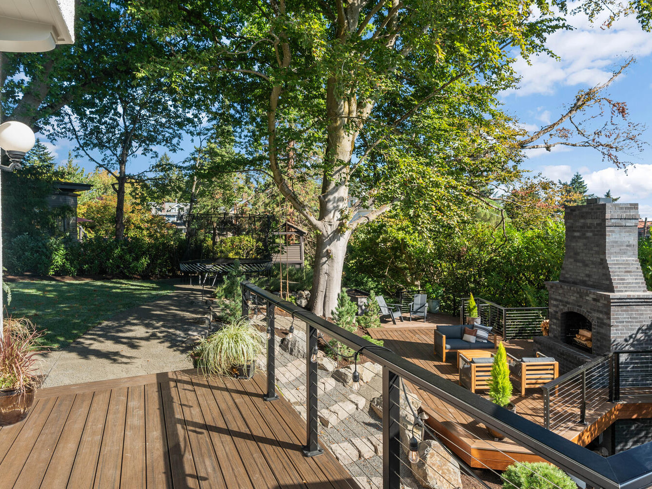 A backyard in Portland, Oregon, features a wooden deck with glass railing and a stylish patio set. A stone outdoor fireplace surrounded by chairs adds warmth. A large tree provides shade amid lush greenery, while the clear, sunny sky completes this serene Northwest retreat.