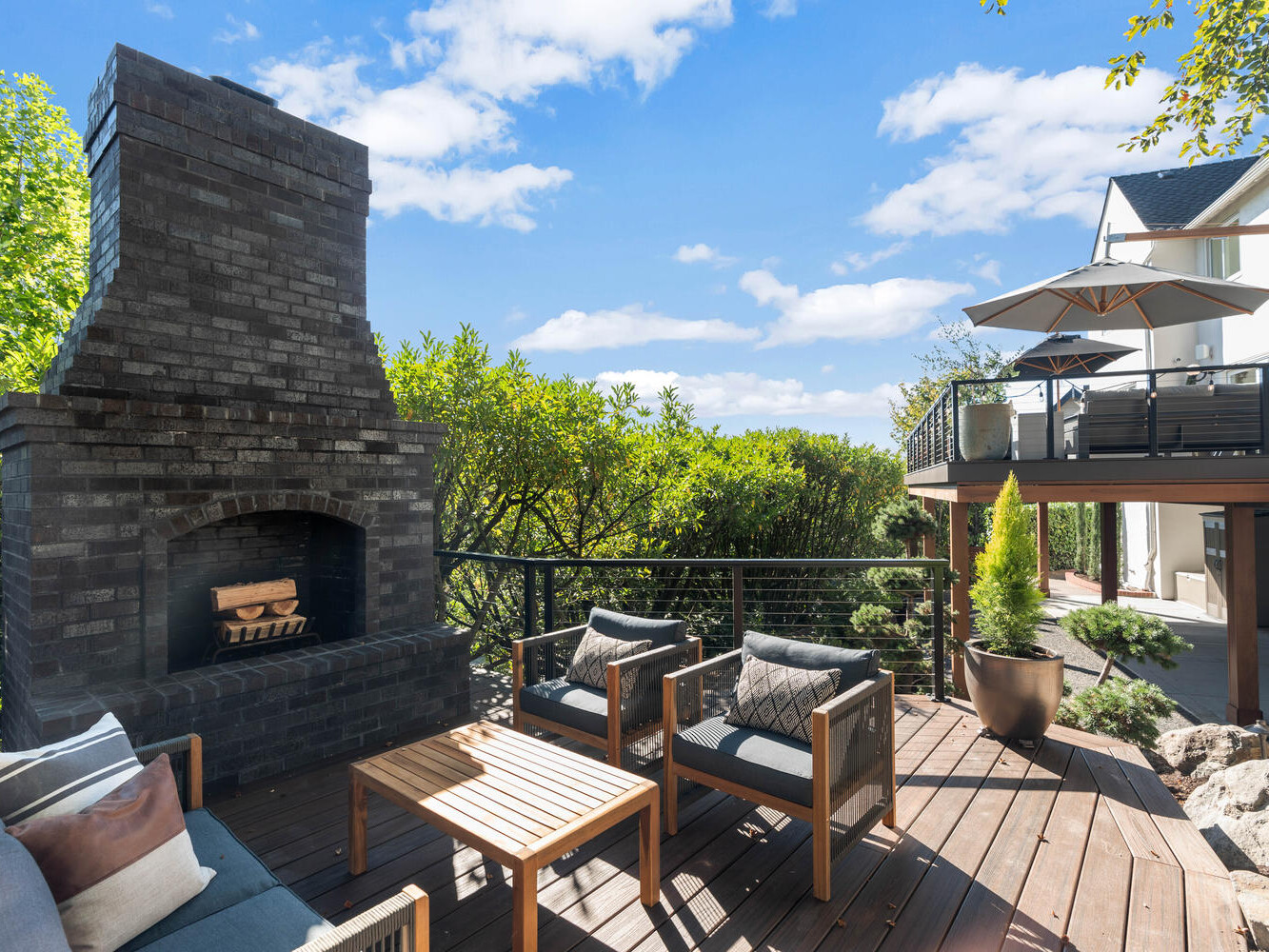 A spacious wooden deck in Portland, Oregon features a large brick fireplace. The seating area includes wooden chairs with cushions and a matching table. Greenery embraces the deck, while a staircase ascends to a second level with an umbrella-shaded seating area.