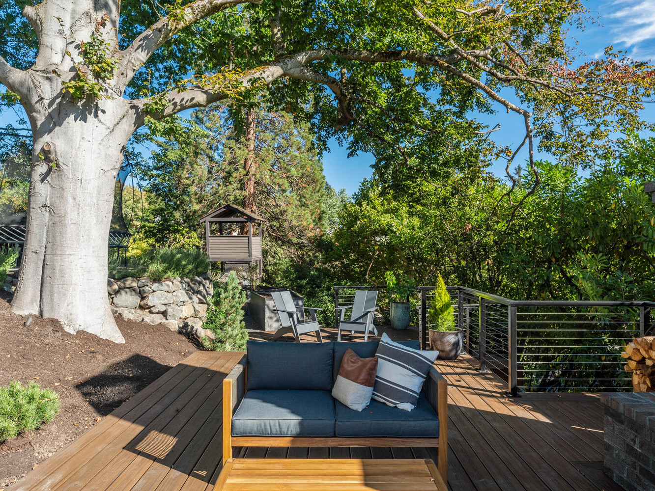 A peaceful Portland, Oregon backyard scene with a wooden deck featuring a gray couch and chairs. A large tree provides shade, while a small wooden playhouse is visible in the background. Lush greenery surrounds the area, creating a tranquil atmosphere.