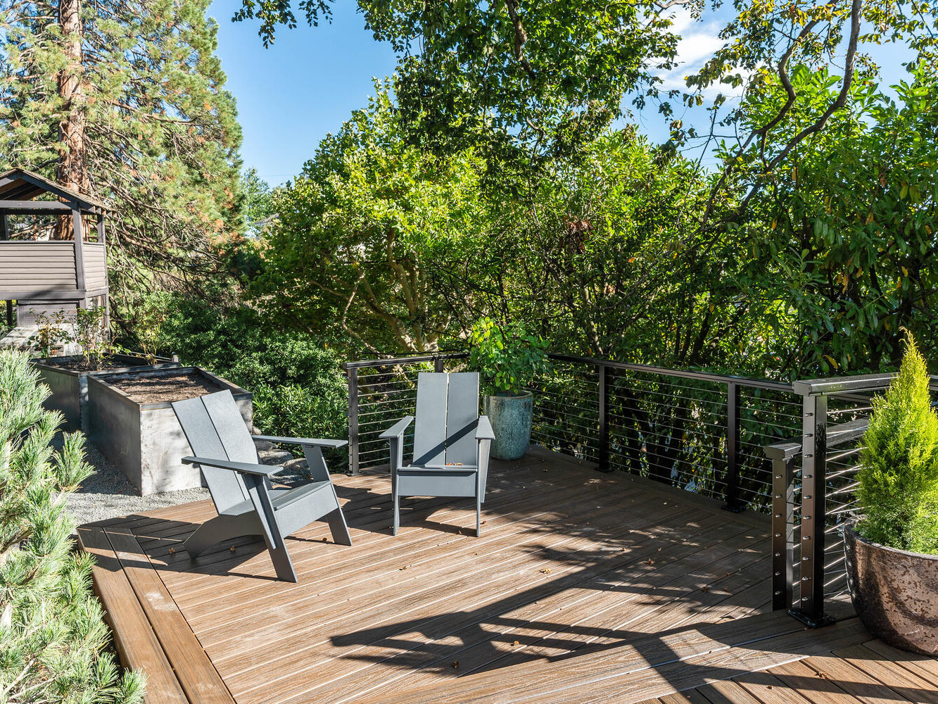 A wooden deck with two gray chairs surrounded by lush green trees captures the serene charm of Portland, Oregon. A planter with a small evergreen enhances the setting, while a small wooden structure, possibly a playhouse, completes this sunlit oasis.