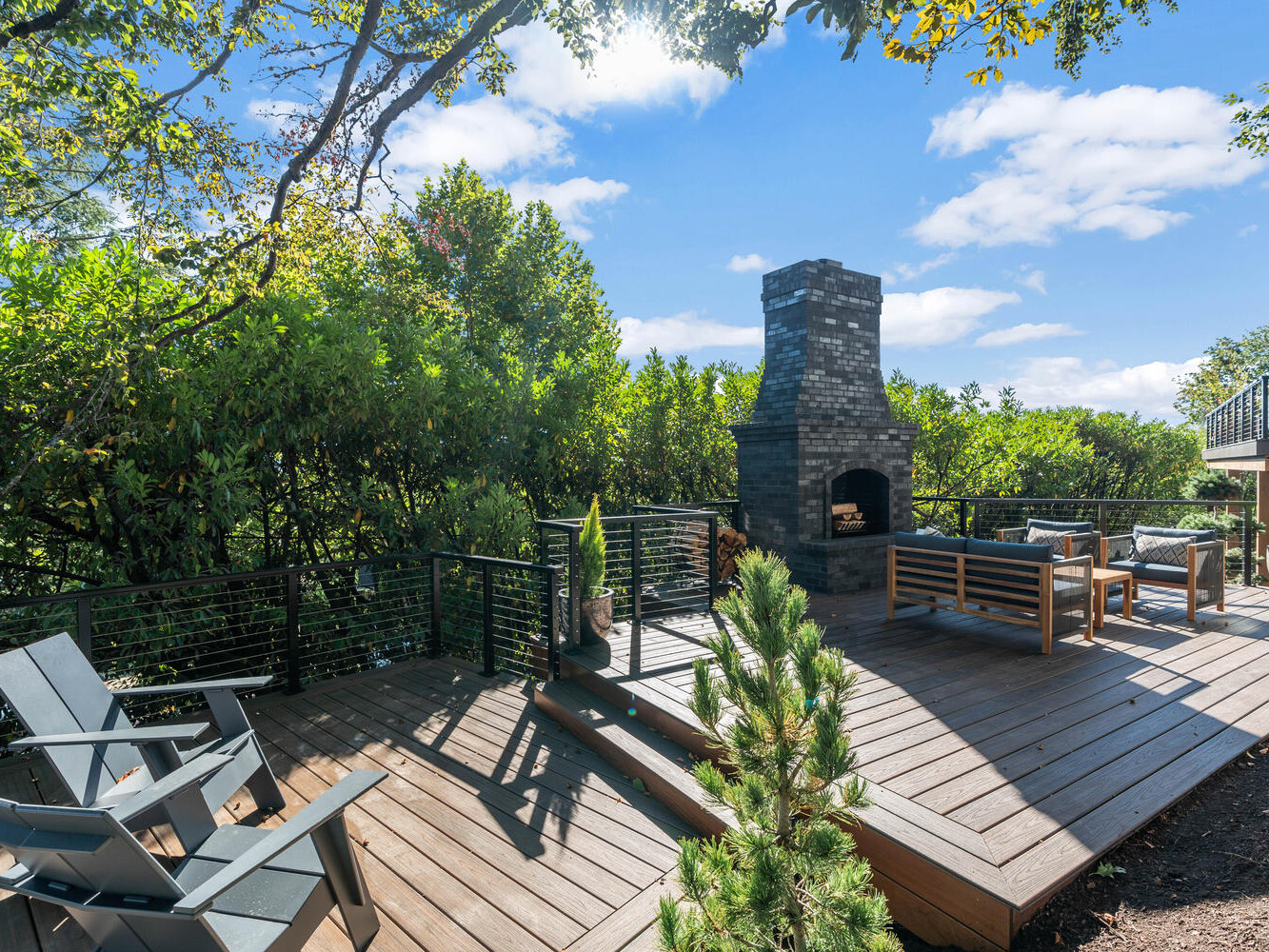 Outdoor patio with wooden decking, nestled in the heart of Portland, Oregon, surrounded by lush greenery. Features a stone fireplace, wooden benches, and modern chairs under a blue sky with fluffy clouds. The serene setting is embraced by trees and decorative plants.