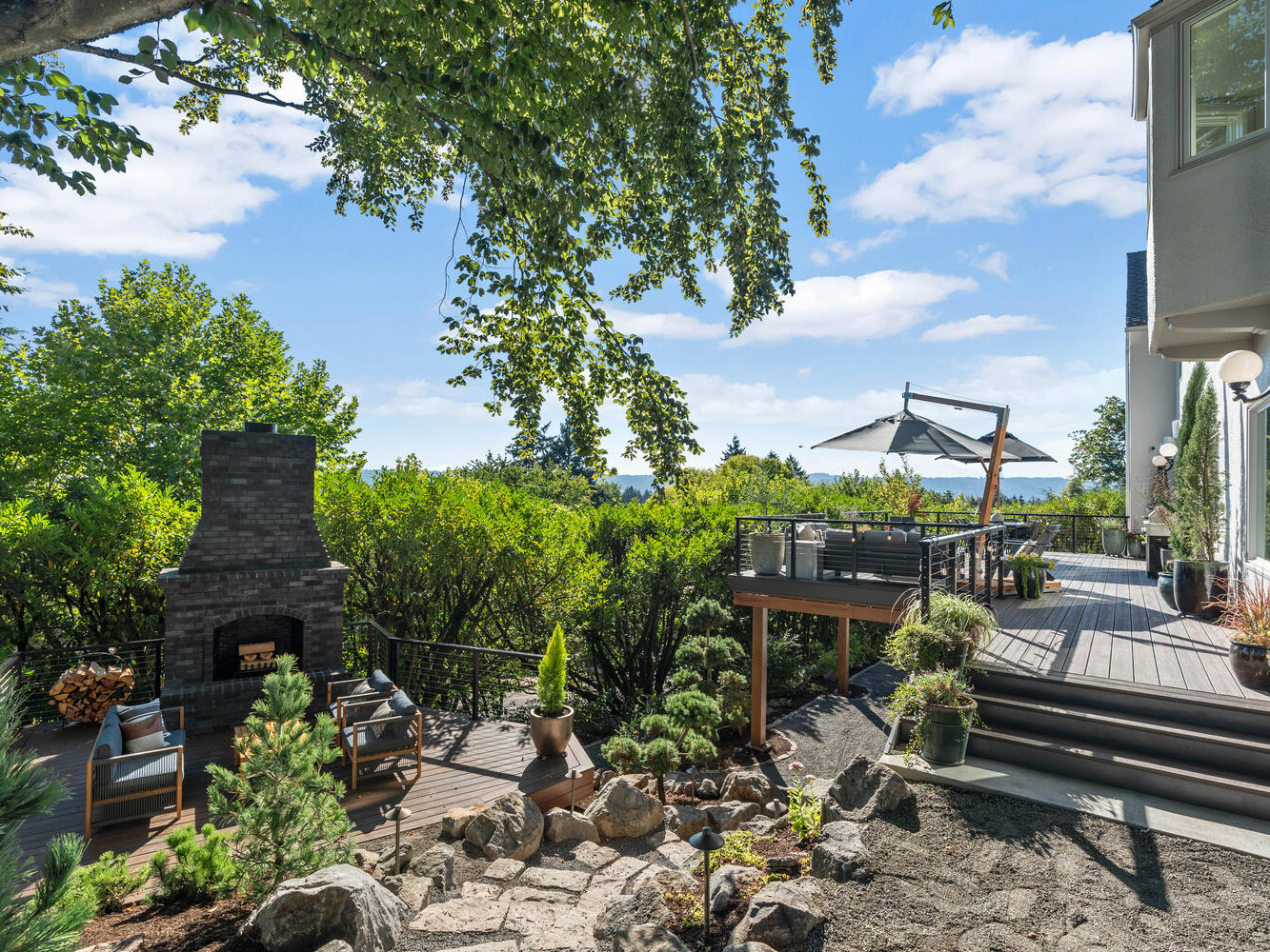 Outdoor seating area with a brick fireplace and two chairs under a large tree, capturing the charm of Portland, Oregon. The adjacent raised deck with an umbrella offers views of distant water and lush greenery. It's a bright, sunny day with scattered clouds gracing the sky.
