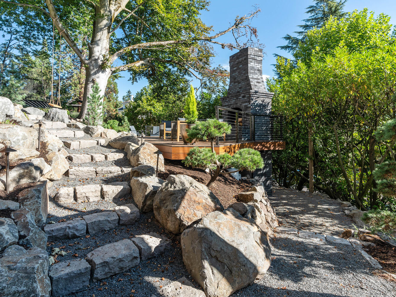 A stone path winds through a landscaped garden with large rocks and trees, leading to a wooden deck in Portland, Oregon. The deck features a stone fireplace and outdoor seating, surrounded by lush greenery under the clear blue sky.