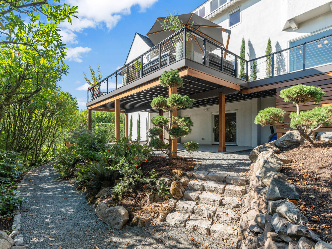 A modern two-story house in Portland, Oregon, boasts a large balcony surrounded by lush greenery and a stone path to the entrance. The balcony features a railing, while the landscaping includes trimmed bushes and trees under the clear blue sky.