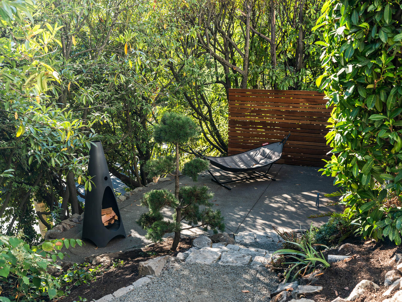 A serene outdoor patio in Portland, Oregon features a hammock surrounded by lush green trees and plants. A modern, black chiminea stands on the stone pavement, adding a cozy touch. Sunlight filters through the foliage, creating a peaceful atmosphere.