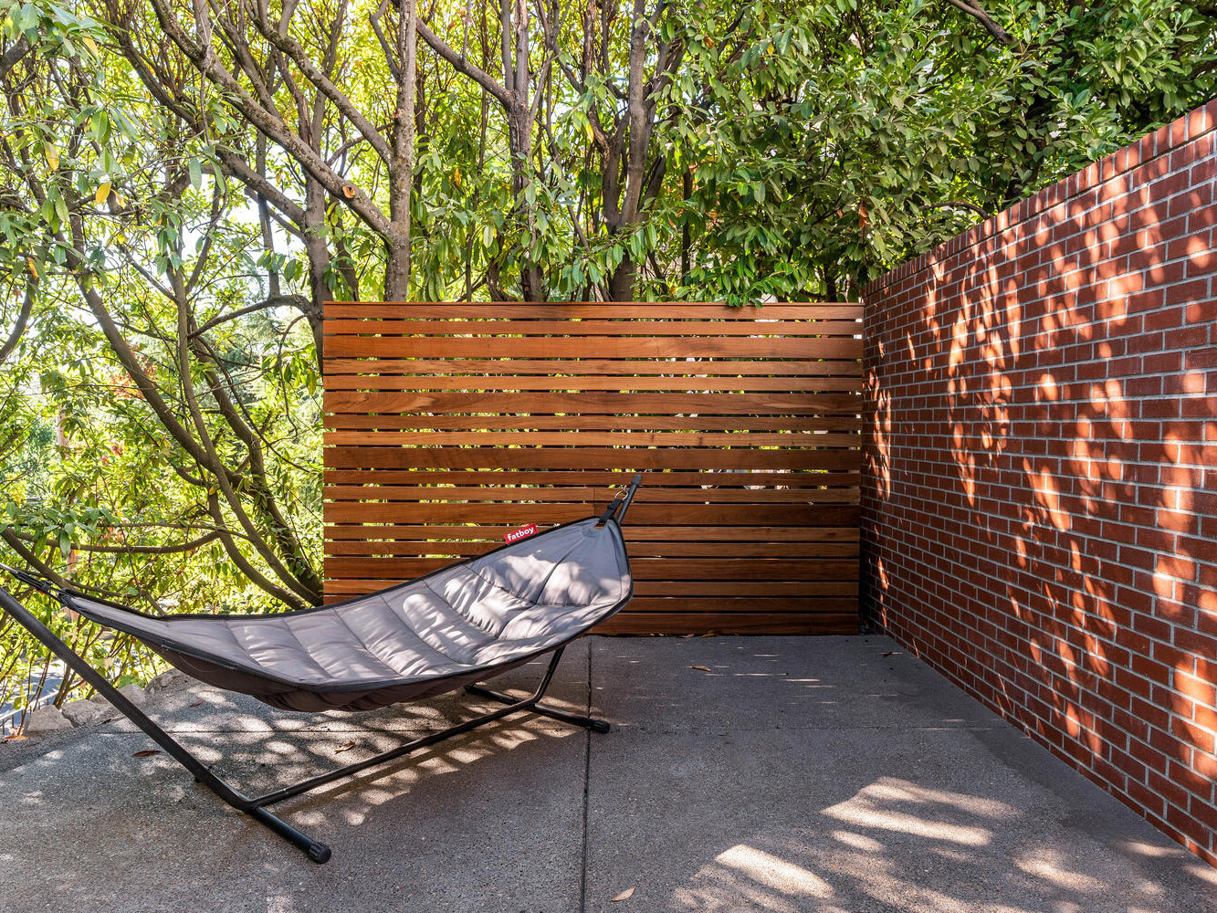 A gray hammock on a metal stand sits on a concrete patio, reminiscent of Portland, Oregon's style. The area is enclosed by a red brick wall on one side and a wooden slat fence on the other. Lush green trees provide shade, filtering sunlight onto the ground.