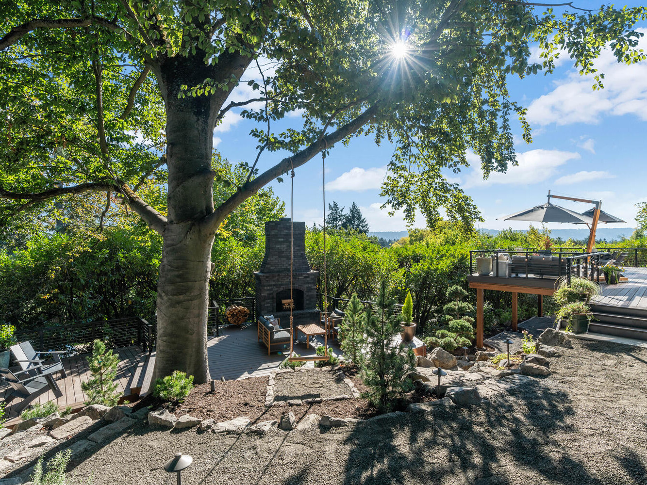 Sunny backyard in Portland, Oregon, with a large tree, outdoor seating, and a fireplace. A deck with an umbrella overlooks lush greenery. The scene boasts clear skies and views of distant water and hills.
