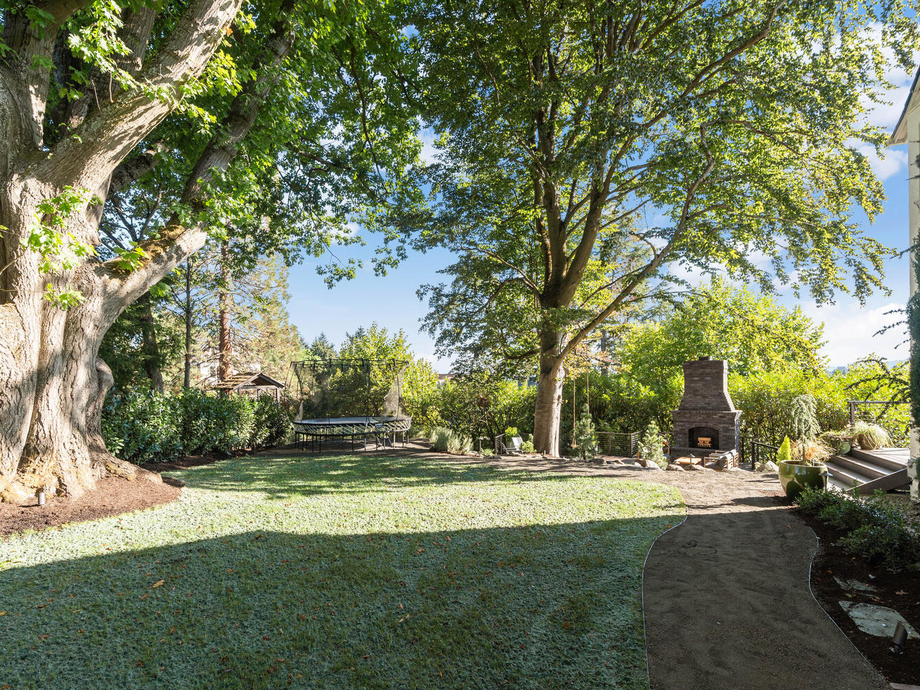 A lush backyard garden in Portland, Oregon features large trees providing shade. The scene includes a neatly manicured lawn, stone pathway, outdoor fireplace, and comfortable seating areas. Sunny weather with a clear blue sky enhances the serene atmosphere.