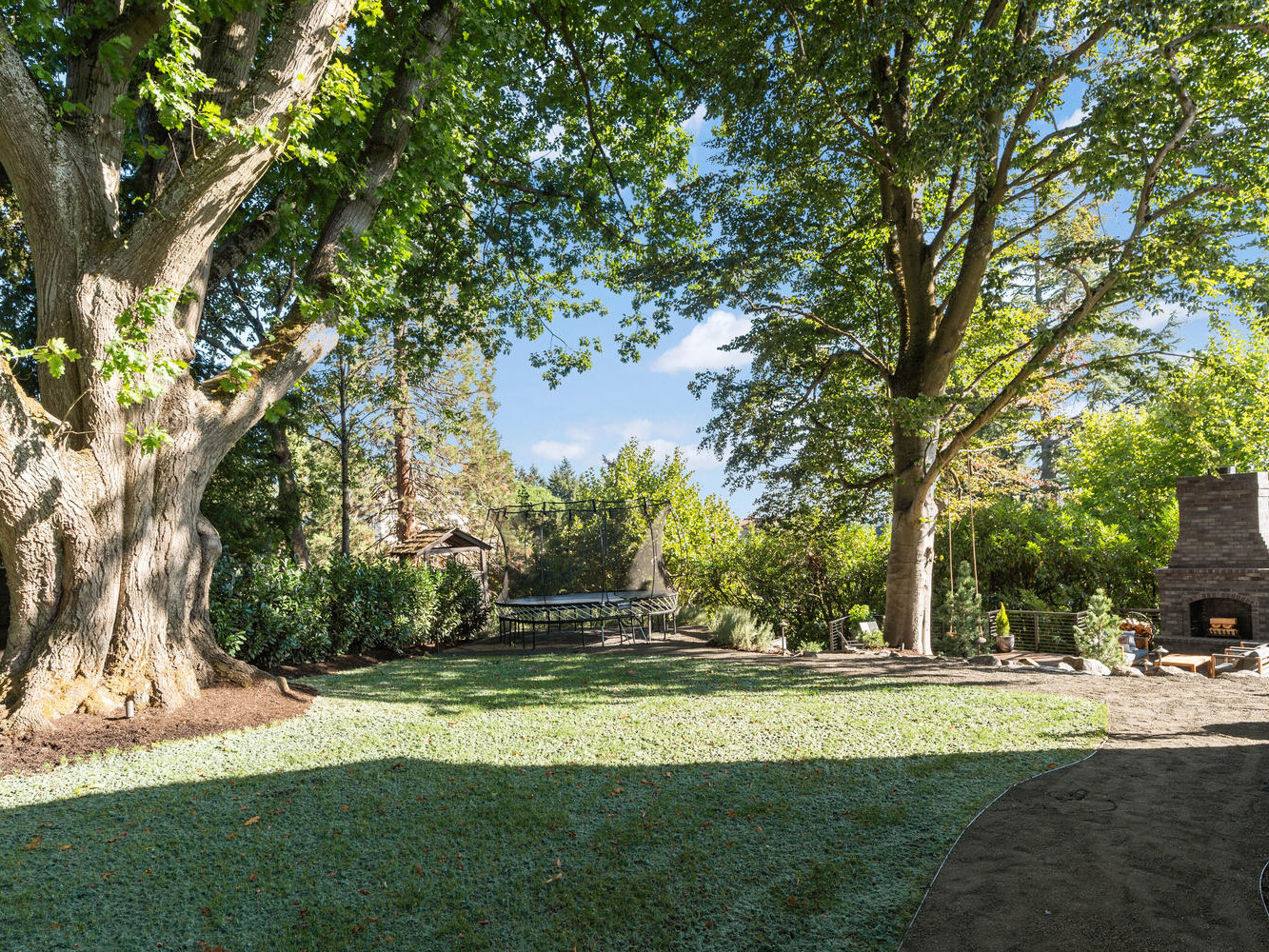 A serene Portland, Oregon backyard features lush green grass, two large trees providing shade, a stone pathway, and an outdoor fireplace. In the background, there's a trampoline surrounded by bushes, creating a peaceful and inviting atmosphere.